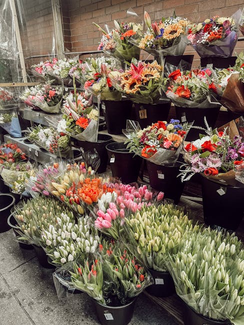 A display of numerous flower bouquets arranged in black buckets and plastic wrappers, featuring a variety of vibrant flowers including pink and white tulips, orange roses, and mixed seasonal blooms. T