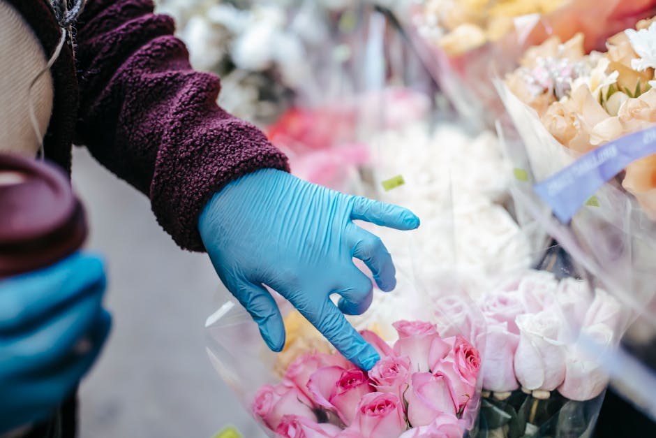 A person's hand wearing a bright blue disposable glove is selecting a bouquet of pink roses wrapped in clear plastic at a flower stall. The bouquet features fresh, pink roses with slightly opened peta