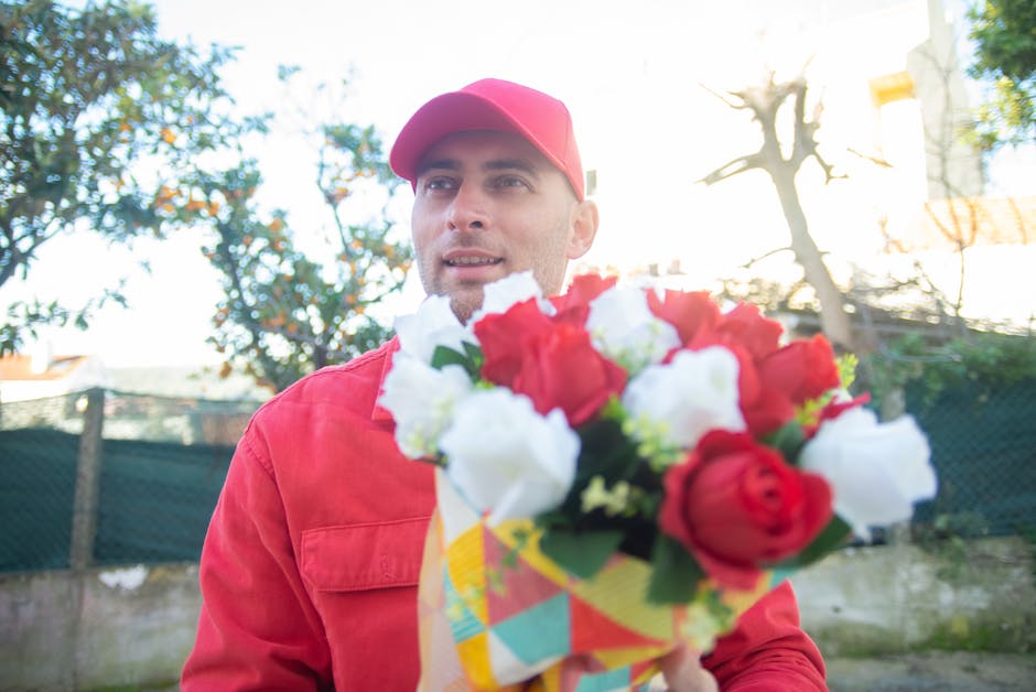 A man wearing a red cap and matching red jacket is holding a vibrant floral bouquet wrapped in colorful paper. The bouquet features red and white roses, with some green foliage visible among the bloss