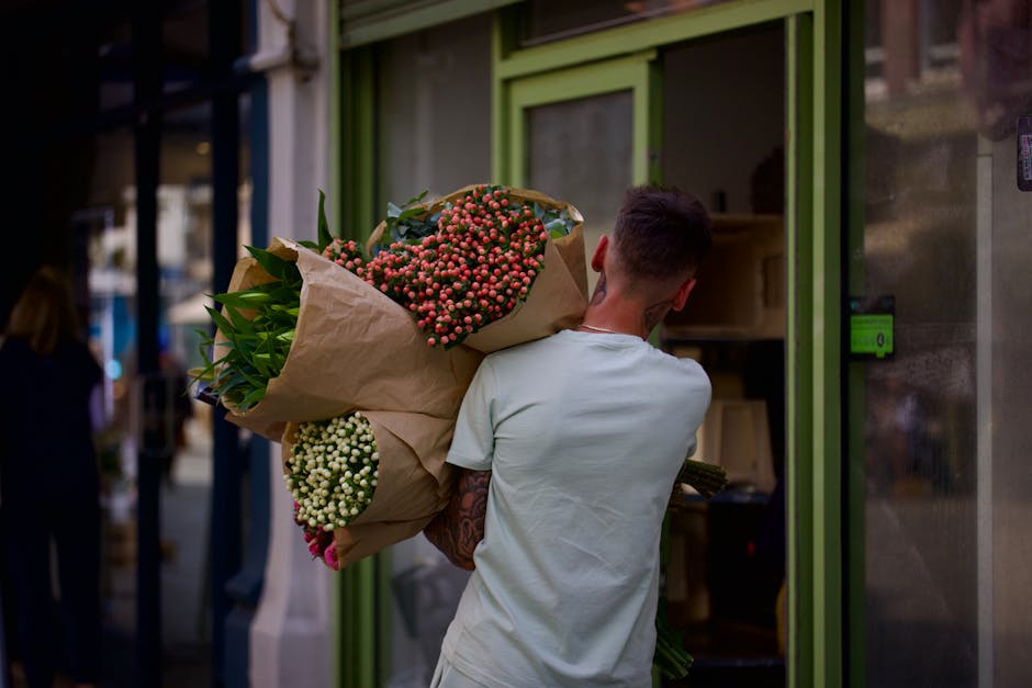 A man with short dark hair, wearing a light grey t-shirt, is carrying a large bouquet of fresh flowers wrapped in brown paper. The bouquet features pink and white blooms, along with green foliage, and