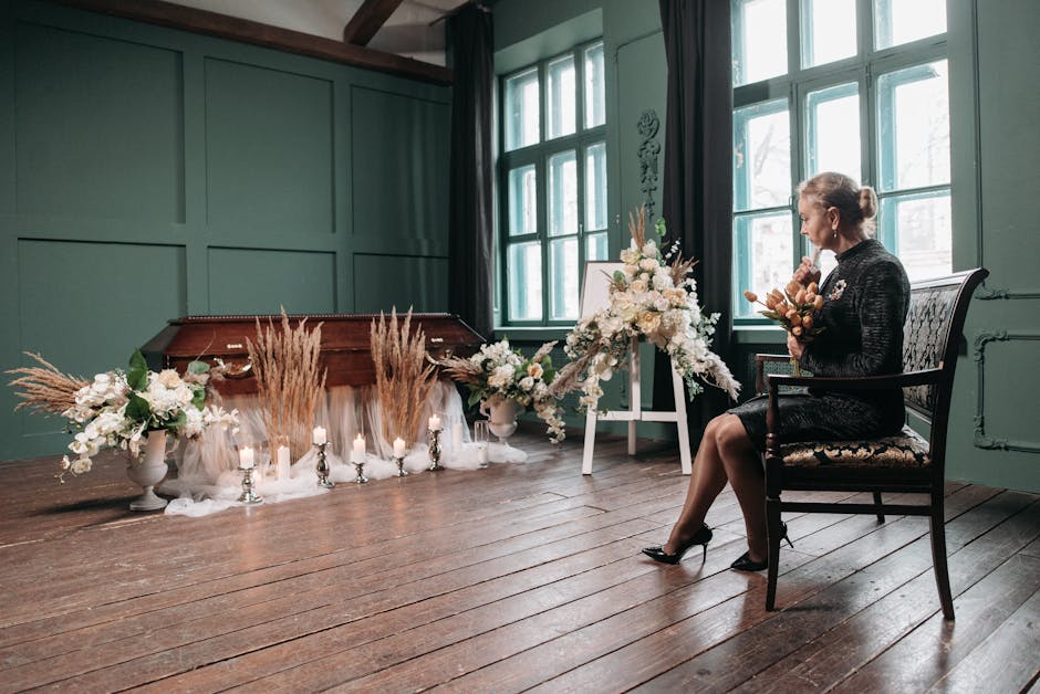 A woman seated on an ornate wooden chair in a softly lit room, holding a small bouquet of dried flowers. In front of her, a floral arrangement on a white easel features predominantly white roses and l