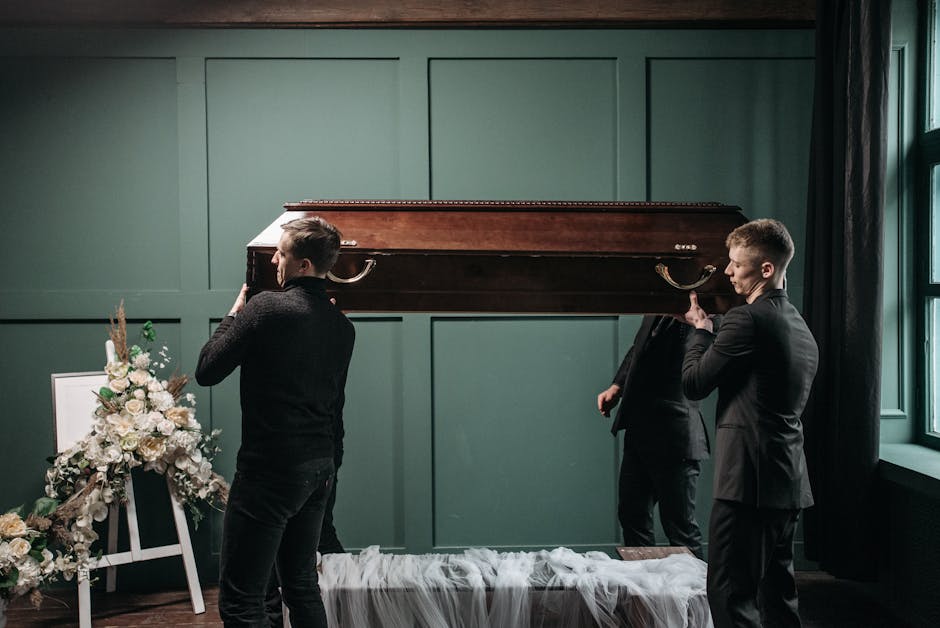 Three men dressed in dark clothing are carrying a closed wooden casket into a room with green paneled walls and large windows. One man is on the left, holding the front of the casket, while another on