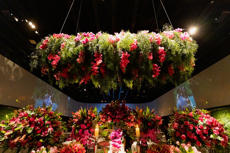 A large, lush floral arrangement hangs overhead, featuring pink and red flowers, including roses and possibly peonies, interspersed with green fir branches, suspended from the ceiling by thin wires in