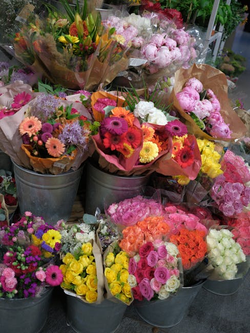 A vibrant display of fresh flower bouquets arranged in metal buckets and wrapped in brown and pink paper, featuring a variety of colorful blooms including gerbera daisies in shades of pink, orange, ye