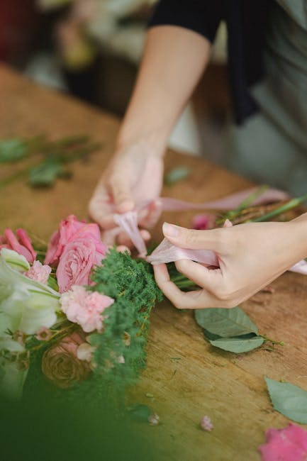 Close-up of a person's hands wrapping a floral bouquet with pale pink ribbon around a arrangement of fresh pink roses, soft green foliage, and small pink flowers. The flowers are arranged on a wooden