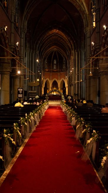 The interior of St John's Church in Pinner, featuring a central aisle covered with a plush red carpet, flanked by dark wooden pews with white floral arrangements and greenery tied with white ribbons.
