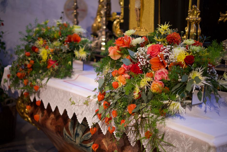 A floral arrangement featuring a vibrant mix of orange, red, pink, white, and yellow flowers, including roses, daisies, and small baby's breath, with lush green foliage and trailing branches. The bouq