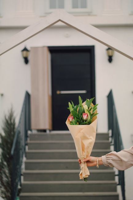 A person extending their arm to present a bouquet of fresh flowers, including pink tulips and green foliage, wrapped in light brown paper, in front of a modern residential entrance. The arrangement is