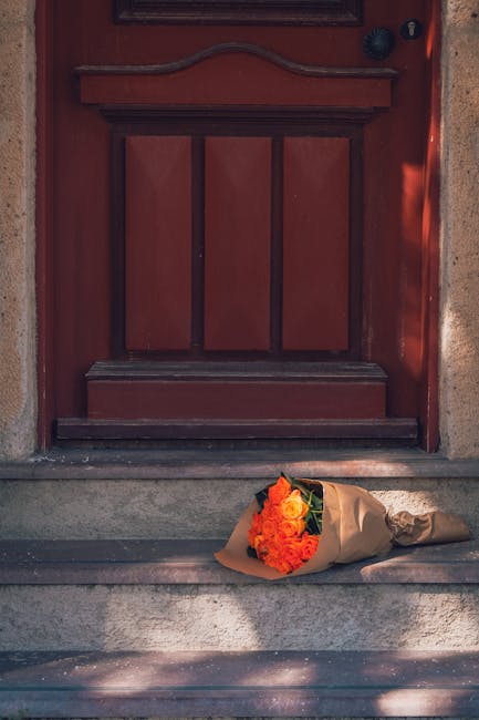 A bouquet of fresh orange roses and mixed flowers wrapped in brown paper rests on stone steps in front of a closed red wooden door. The bouquet features vibrant, slightly open petals with visible text