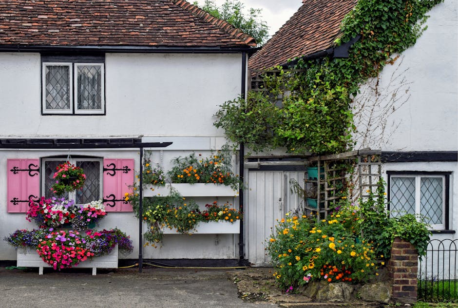 The image depicts a charming white cottage with a red-tiled roof, featuring decorative black window frames with diamond-patterned panes. The facade is adorned with vibrant floral displays, including a